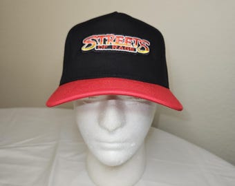 Puede incluir: Una gorra de béisbol negra con visera roja que presenta un parche bordado en la parte delantera. El parche muestra el texto "STREETS OF RAGE" en una fuente estilizada con degradado amarillo, naranja y rojo con borde negro.