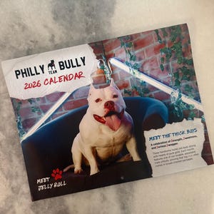 May include: A 2026 calendar featuring a white pit bull with a red collar, sitting on a dark blue chair. The calendar has the text "PHILLY BULLY TEAM" and "MEET JELLY ROLL". The text "MEET THE THICK BOYS" is also visible.