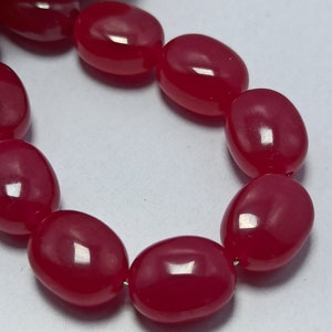 May include: A strand of smooth, polished, oval-shaped beads in a vibrant, translucent cherry red colour. The beads are strung together on a thin wire, showcasing a glossy finish that reflects light across their rounded, uniform surfaces.