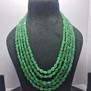 May include: A multi-strand necklace featuring oval-shaped green beads. The necklace is displayed on a black bust, showcasing the cascading layers of the beads. The beads have a polished finish, reflecting light and adding a touch of elegance.