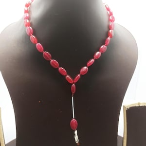 May include: A red beaded necklace displayed on a black mannequin. The necklace features oval-shaped beads and a silver-colored bar pendant with a small charm. The beads are a deep red color, and the necklace is designed to be worn around the neck.