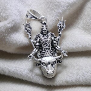 May include: A silver pendant depicting a deity with multiple arms, holding trident-like objects. The deity is standing on a bull's head. The pendant has a loop for a chain. The pendant is silver in colour.