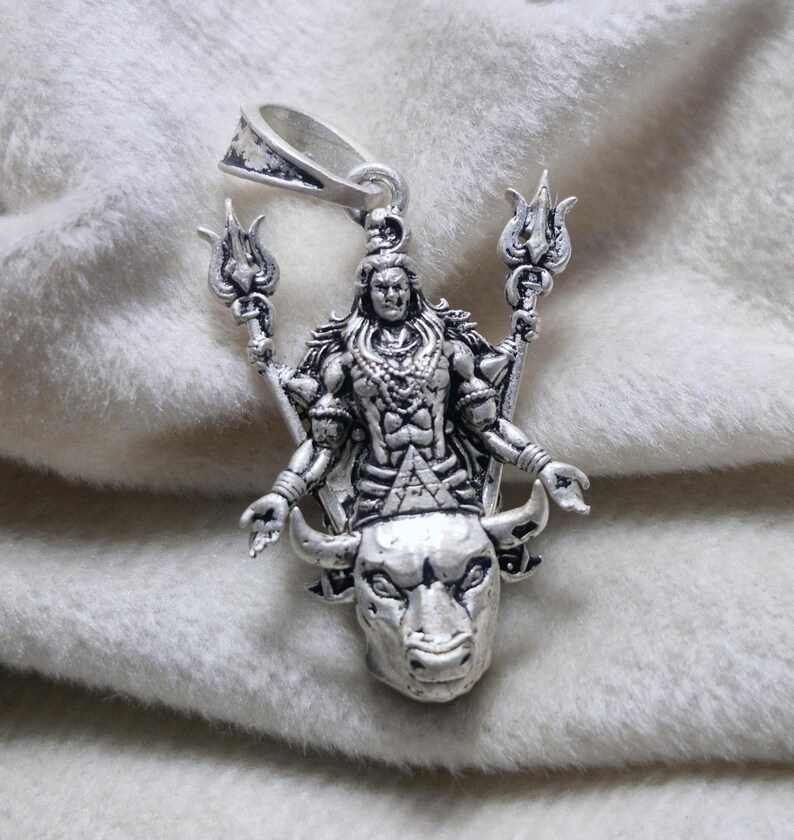 May include: A silver pendant depicting a deity with multiple arms, holding trident-like objects. The deity is standing on a bull's head. The pendant has a loop for a chain. The pendant is silver in colour.