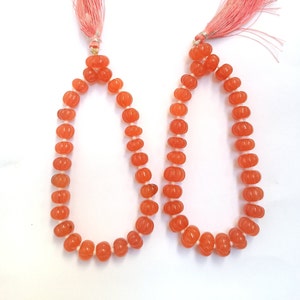 May include: Two strands of orange gemstone beads with a pumpkin-like shape. Each strand is strung on a white cord and finished with a peach-colored tassel. The beads have a glossy finish and are arranged in a symmetrical pattern.