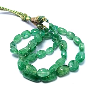 May include: A necklace of polished, oval-shaped emerald beads in varying shades of green. The beads are strung together with a gold-coloured cord and a green, braided tassel. The background is white.