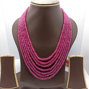 Peut inclure: Un collier à plusieurs rangs composé de petites perles rondes de couleur magenta. Le collier est présenté sur un buste à bijoux marron, mettant en valeur son design superposé. Les perles ont une taille et une forme uniformes.