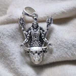May include: Silver pendant depicting a deity with multiple arms, holding trident-like objects. The pendant is mounted on a bull's head, with intricate detailing. It has a loop at the top for a chain. The pendant is likely made of silver.