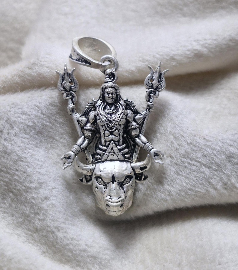 May include: Silver pendant depicting a deity with multiple arms, holding trident-like objects. The pendant is mounted on a bull's head, with intricate detailing. It has a loop at the top for a chain. The pendant is likely made of silver.