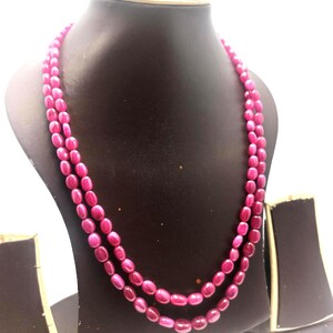 Può includere: Una collana a due fili con perline ovali rosa intenso. La collana è esposta su un busto di gioielli nero su sfondo bianco. Le perline hanno una finitura lucida che riflette la luce.