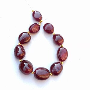 Op de afbeelding: Snoer van gepolijste, ovaalvormige kralen in een diepe bordeauxrode kleur. De kralen zijn geregen met goudkleurige afstandhouders, geschikt voor een ketting of armband. De kralen hebben een glanzende afwerking.