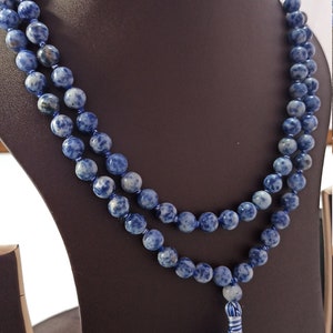 May include: A beaded necklace with blue and white speckled stones. The necklace is displayed on a brown mannequin. A blue and white tassel hangs from the bottom of the necklace. The necklace is made of multiple strands of beads.