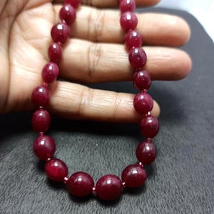 Puede incluir: Un collar de cuentas de piedras preciosas pulidas de color rojo intenso. Las cuentas están ensartadas, mostrando una variedad de formas, desde redondas hasta ligeramente ovaladas. El collar se exhibe sobre un fondo neutro, resaltando el rico color y brillo de las piedras preciosas.