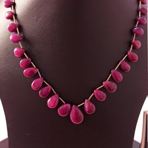 May include: A necklace featuring a single strand of deep pink, teardrop-shaped gemstone beads strung on a thin gold-colored wire. The beads are uniform in shape and size, arranged symmetrically along the length of the necklace chain.