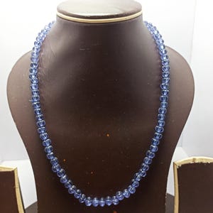 Könnte beinhalten: Eine Halskette aus kleinen, runden, hellblauen Perlen. Die Perlen sind zu einer Halskette aufgereiht. Die Halskette wird auf einer braunen Schmuckbüste vor weißem Hintergrund präsentiert. Die Perlen haben ein durchscheinendes Aussehen.