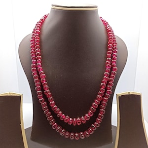 May include: A layered necklace with red beads is displayed on a brown jewelry bust. The beads are small, round, and a deep red color. The necklace is draped in two strands, showcasing the design and color.