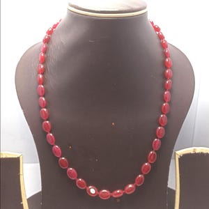 Puede incluir: Un collar de cuentas rojas expuesto sobre un maniquí negro. El collar presenta cuentas de forma ovalada en un tono rojo intenso. Las cuentas están ensartadas, creando un bucle continuo. El collar se presenta sobre un fondo blanco.