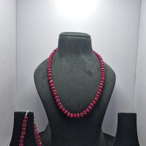 Può includere: Un set di collana e bracciale con perline rosso rubino, esposti su supporti neri. La collana è drappeggiata su un manichino, mentre il bracciale poggia su un supporto più piccolo. Il bracciale presenta una chiusura color oro.