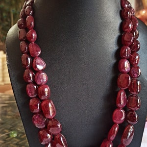 May include: A double-strand necklace of large, polished, oval ruby beads. The beads are a deep red hue and are strung together. The necklace is displayed on a black jewellery bust.