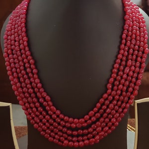 May include: A multi-strand necklace featuring five layers of smooth, polished, oval-shaped red beads. The beads are uniform in size and color, strung together to create a cascading effect that rests against the dark display form.