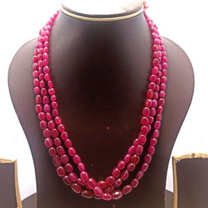 Op de afbeelding: Een meerstrengs ketting met ovale, dieprode kralen. De ketting wordt gepresenteerd op een zwarte mannequin, die het gelaagde ontwerp en de rijke kleur van de edelstenen benadrukt. De kralen hebben een gepolijste afwerking.