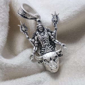 May include: A detailed silver pendant depicting a deity figure with multiple arms, holding trident-like objects. The figure is adorned with intricate jewellery and stands above a bull's head. The pendant has a textured finish and a loop for a chain.