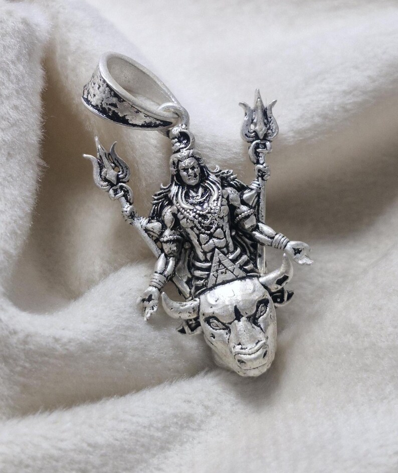 May include: A detailed silver pendant depicting a deity figure with multiple arms, holding trident-like objects. The figure is adorned with intricate jewellery and stands above a bull's head. The pendant has a textured finish and a loop for a chain.