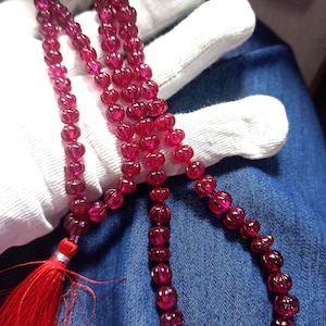 May include: A strand of deep red gemstone beads, possibly ruby, with a tassel. The beads are strung together, forming a necklace. The necklace is held by a gloved hand, against a denim background.