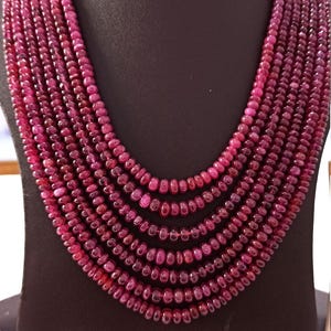 Op de afbeelding: Een meerstrengs ketting bestaande uit kleine, ronde, dieprode kralen. De ketting wordt getoond op een zwarte mannequin buste, die de trapsgewijze lagen van de sieraden benadrukt. De kralen hebben een gepolijste afwerking, die het licht reflecteert.