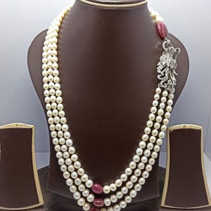 Puede incluir: Un collar de perlas de tres hilos con un cierre decorativo plateado con un diseño de pájaro. El collar tiene tres hilos de perlas blancas, intercaladas con piedras preciosas rojas. El collar se exhibe en un busto de joyería marrón.