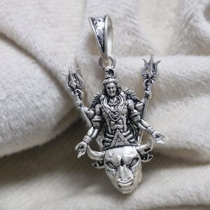 May include: A silver pendant depicting a deity with multiple arms, holding trident-like objects. The pendant is mounted on a bull's head, with intricate detailing. It has a loop for a chain. The pendant is made of silver.