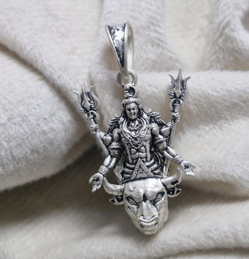 May include: A silver pendant depicting a deity with multiple arms, holding trident-like objects. The pendant is mounted on a bull's head, with intricate detailing. It has a loop for a chain. The pendant is made of silver.