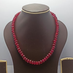 Op de afbeelding: Een rode edelsteen kralenketting op een bruine displaystandaard. De ketting is gemaakt van ronde, gefacetteerde kralen.
