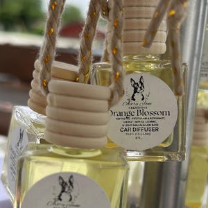 May include: Clear glass car air fresheners filled with a yellow liquid, labeled "Orange Blossom." Each bottle has a wooden cap and is suspended by a gold-flecked rope. The label includes the text "100% Organic" and "8ml."