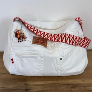 May include: White denim shoulder bag with a red and white patterned strap. The bag features a camel charm and a Levi Strauss & Co. label. The bag has a zip closure and two pockets.