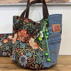 May include: A tote bag constructed from a combination of dark fabric featuring a multi-coloured bird and floral tapestry pattern and blue denim fabric with a Levi Strauss & Co. leather patch. The bag has brown handles and a green beaded charm.
