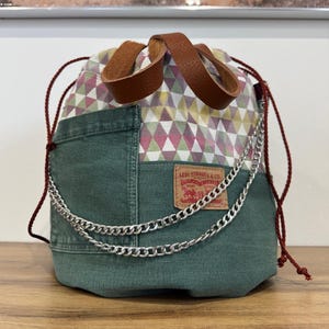 Könnte beinhalten: Ein grüner Jeans-Beutel mit Kordelzug, geometrischem Muster oben und braunen Ledergriffen. Eine silberne Kette und ein Levi Strauss & Co. Etikett sorgen für Details. Die Tasche ist etwa 25 cm hoch.