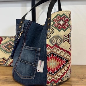 May include: A denim tote bag with black leather handles and a decorative patterned fabric panel. The bag features a front pocket and a blue beaded charm. The patterned fabric has red, green, and beige geometric designs.
