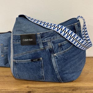 May include: A denim shoulder bag with a blue and white patterned strap. The bag is made from recycled jeans and features the Calvin Klein logo on a black patch. The bag has a curved shape and a pocket on the front.