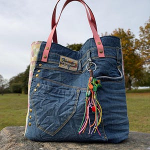 May include: A denim tote bag featuring pink leather handles, a Desigual brand patch, and a multi-colored tassel charm. The bag includes external pockets, metal rivets, and side panels with a geometric pattern in pastel shades of yellow, pink, and green.