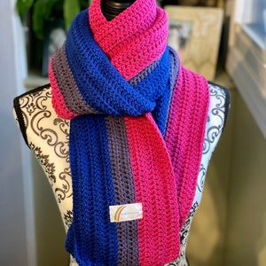 Może przedstawiać: Ręcznie robiona szydełkowa szal w odcieniach różu, niebieskiego i szarości, narzucona na manekina. Szal ma wzór w paski i małą etykietę z tęczą i tekstem. Przytulny dodatek.