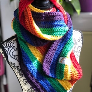 Könnte beinhalten: Ein regenbogenfarbener gestrickter Schal mit einem kleinen Regenbogen-Etikett. Der Schal ist gefaltet und über einen schwarzen Mannequin-Busen gelegt. Der Text "Style A" ist in der oberen rechten Ecke des Bildes sichtbar.