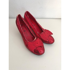 May include: A pair of bright red sequined pumps featuring a rounded toe and a decorative bow on the front. The interior of the shoes is lined in red material with visible text printed on the insole, and the exterior is covered in small red sequins.