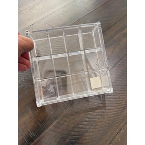 May include: A clear acrylic organizer box featuring twelve individual square compartments arranged in a grid pattern. The transparent container has a rectangular frame and a small rectangular sticker label on the bottom right corner.