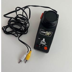 May include: Black Atari paddle controller with a red button and a black dial. The controller has the word "paddle" and the Atari logo printed on it. It includes a black cable with white and yellow RCA connectors.