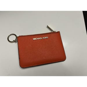 May include: An orange Michael Kors cardholder with a silver zipper and key ring. The cardholder has the brand name in gold lettering. The item is rectangular and made of a textured material.
