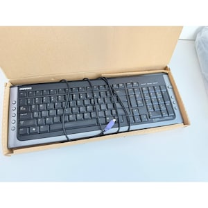 May include: A Compaq brand computer keyboard with a black and gray design. The keyboard is inside a cardboard box and has a wired connection with a purple connector. The keyboard has a standard QWERTY layout.