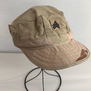 May include: Tan military-style cap with a short brim and a square patch featuring black chevrons. The cap has a structured design with a slightly distressed look, and a camouflage pattern on the brim.