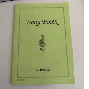 May include: A light green songbook with the title "Song BooK" in black font. A treble clef graphic is centered below the title. The brand name "CASIO" is printed at the bottom. The book has a decorative border.