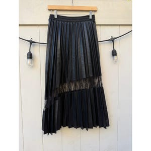May include: A black pleated midi skirt featuring a smooth, faux leather finish and a horizontal band of intricate black lace detailing across the lower section. The skirt has a wide, elasticized waistband and hangs from a wooden clothes hanger.