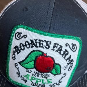 Może przedstawiać: Czarna i biała czapka trucker z naszywką haftowaną na zielono i biało, na której widnieje napis "Rooneys Farm" oraz czerwone jabłko z zielonymi liśćmi. Naszywka zawiera również napis "Old Fashioned 100% Pure Apple Wine Sauce."
